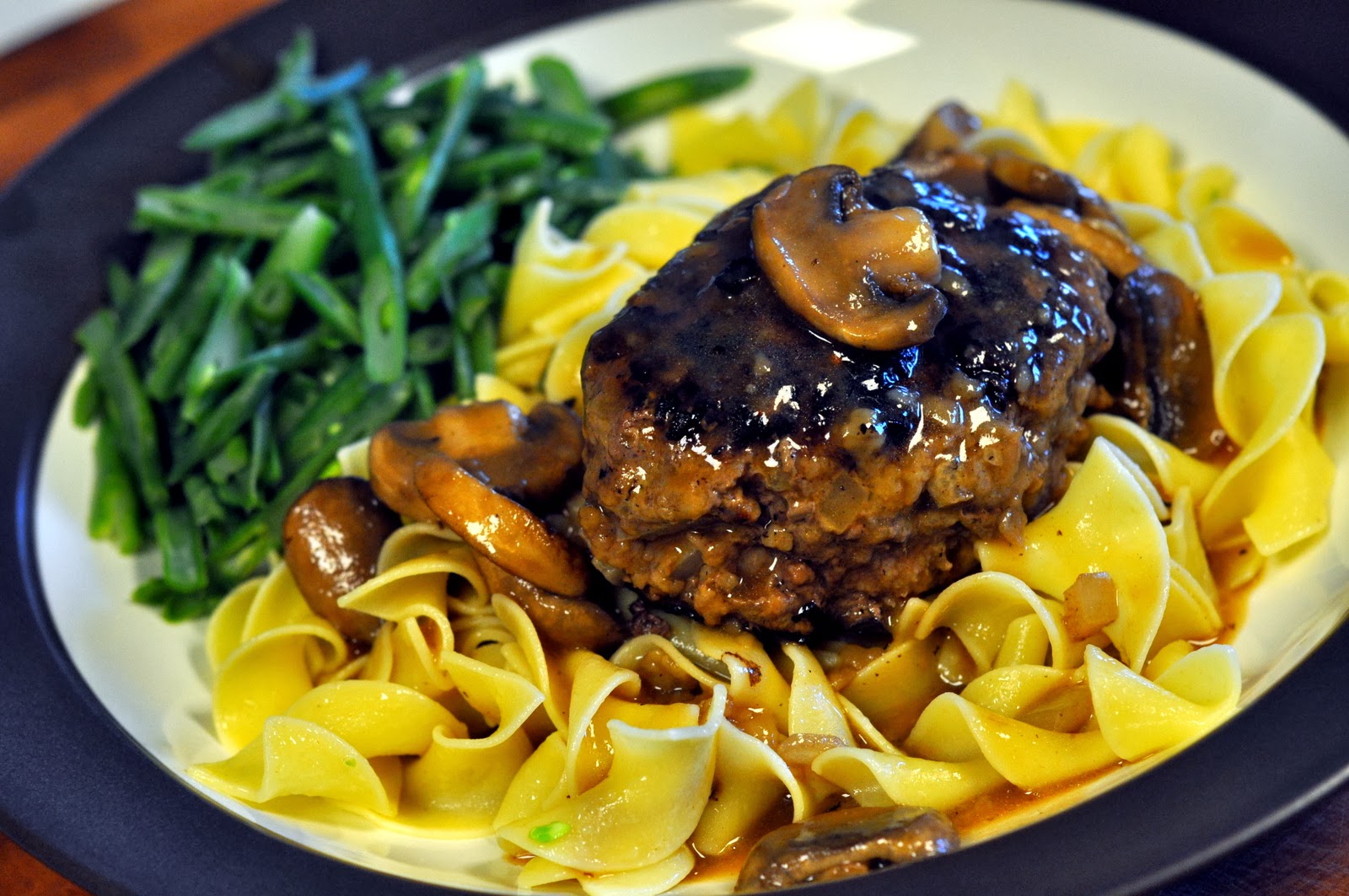Salisbury steak with mushrooms over wide egg noodles and served with green beans. Food is on a blue and white plate.