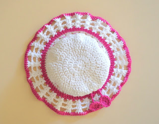 Above-view of the hat. The crown is white with pink borders where the crown meets the brim and at the end of the brim. The body of the brim is a lacy crossed stitch pattern.  The two discs that decorate the ends of the drawstring can also be seen.