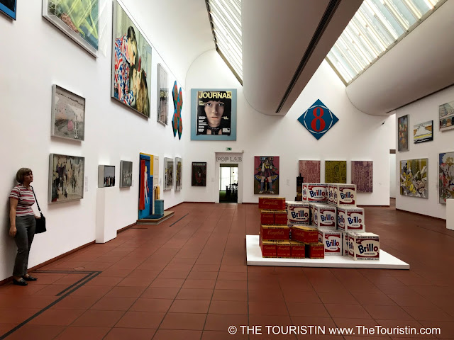 A large hall decorated with paintings and art pieces that are replicas of washing powder packages, with a lifelike sculpture of a woman looking at the art.