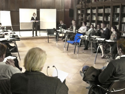 Ein dunkel getäfelter Tagungsraum mit Seminarteilnehmern und Vortragender. Aus dem in Naturtönen gehalten Bild sticht ein hellblauer Stuhl hervor.