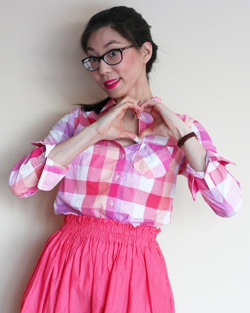 [Image of a tan skin bespectacled Asian woman wearing a pink plaid button down tucked into an elastic waist A-line skirt she made years ago. Dark brown hair in a French braid. Her hands are making a heart-shape as she smiles at you.]