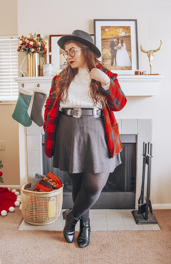A woman with brown wavy hair wearing a black wide brim fedora, white pleated Victorian inspired blouse, a red and black buffalo plaid coat, a black short skater skirt, a black and silver western belt, black tights and black heeled ankle Chelsea boots