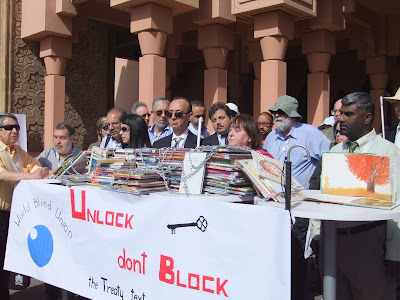 Action de l'Union mondiale des Aveugles à Marrakech : sur une table, des piles de livres sont couverts de chaines. Sur le panneau disposé en dessous, il est écrit "Unlock, don't block the Treaty text."