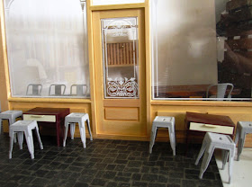 Mock up of the front of a modern dolls' house miniature cafe, showing the shop front and several tables and unpainted cafe chairs.