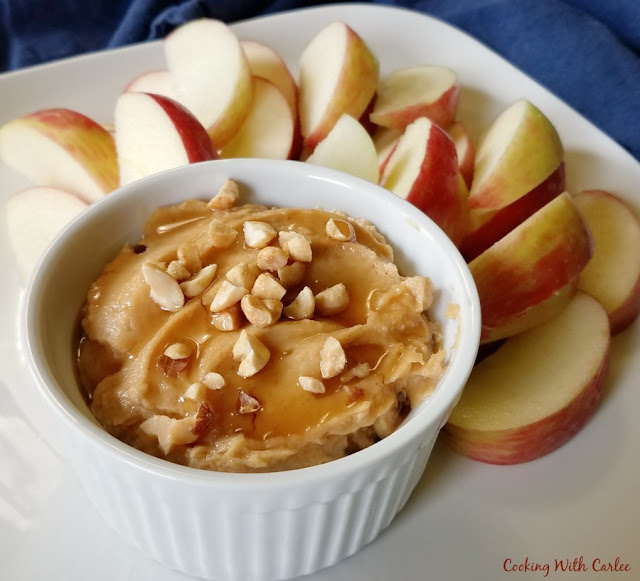 This fun dip is perfect for snacking. It just has a few simple ingredients and comes together in not time at all. The best part is all of the ingredients are wholesome as well. Perfect after school snack or fun for a party! close up of ramekin filled with peanut butter yogurt dip with chopped peanuts and drizzle of honey on top and sliced apples on plate