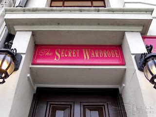 A photograph of a wooden board with a frame around the edged, painted in red with gold vinyl letters 'The Secret Wardrobe' in script and decorative fonts mounted on the entrance above the shop door.
