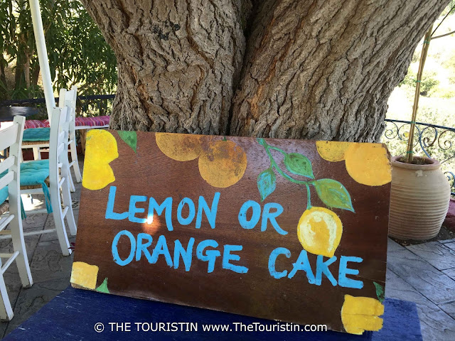 Travel guide to Lindos in Greece. Wooden sign on a blue table under a tree, promoting Lemon or Orange Cake