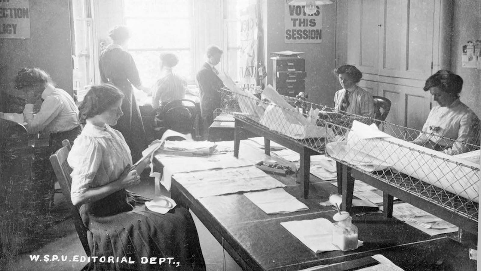 Women in the editorial department of Clement's Inn, The Strand, London. That week's edition of 'Votes for Women' is being cut and pasted by the young woman volunteers at the 'making-up table.' All the woman have long hair tied up in loose buns. Most of the woman wear white blouses and full skirts — two wear neck ties. 1911