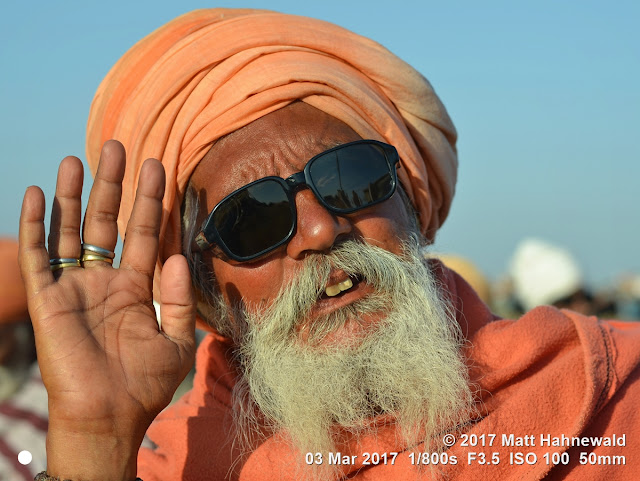 matt hahnewald photography; facing the world; character; head; face; sunglasses; facial expression; beard; teeth; full beard; ring; hand; turban; blessing; greeting; raised right hand; consent; rapport; respect; traveling; spiritual; religious; traditional; cultural; holy; hinduism; temple; sadhu; guru; baba; roadside; rukshmani devi; dwarka; gujarat; western india; asian; indian; person; male; adult; elderly; old; man; image; picture; photo; face perception; physiognomy; nikon d3100; nikkor af-s 50mm f/1.8g; prime lens; 50mm lens; nifty fifty; 4x3; horizontal; street; portrait; closeup; head shot; seven-eighths view; orange; outdoor; sunlight; morning sun; color; posing; authentic