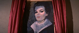 Foto de cena do filme. Um quadro pindurado na parede por trás de cortinas vermelhas. As cortinas estão abertas, revelando o retrato de Barbara Steele. Seu olhar oblíquo parece nos observar do alto, os olhos bem delineados têm iluminação que os destacam. O retrato é bastante fiel e tem um tom levemente azulado. A expressão no rosto dela é quase de superioridade. Ela usa os cabelos negros presos, mas a franja longa cai levemente sobre a orelha semi-aparente. sobre uma gola alta de estruturada branca, outra gola alta em "v", de um casaco ou vestido escuro.