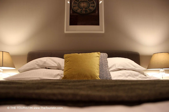 A photo of a clock over a large bed with white yellow and black and white cushions and duvet covers and a grey-green blanket with lamps on both sides.