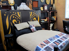 One-twelfth scale miniature scene, with a bed dressed in linen and black with throw rug and cushion printed with letter As, in front of a false wall.