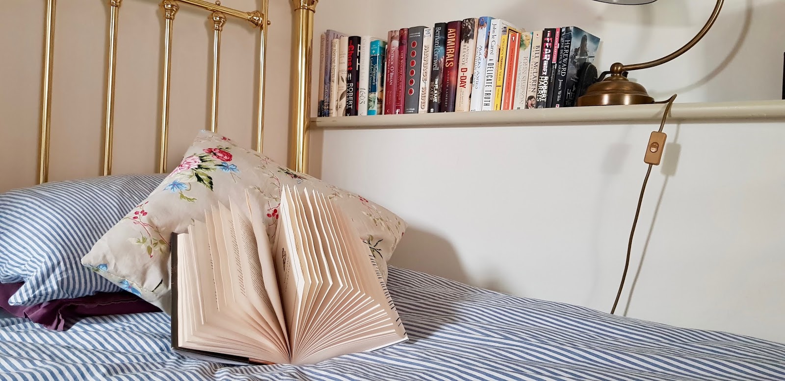 n open book rests on a pillow on a bed with a blue stripe duvet cover, with a shelf of books in the background