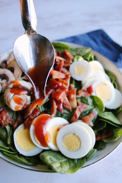 Spinach salad is a favorite for good reason. It is loaded with yummy toppings that work so well with the homemade dressing and spinach base. Serve it as a side or make a meal of it! drizzle spoon drizzling red dressing over bowl of spinach salad with eggs bacon and mushrooms