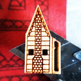 Side of a 'Very small dollshouse' displayed on the tip of a cutting knife, and showing the details of windows, siding and chimney laser-cut into it.