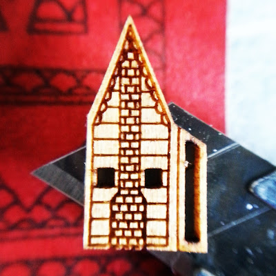 Side of a 'Very small dollshouse' displayed on the tip of a cutting knife, and showing the details of windows, siding and chimney laser-cut into it.