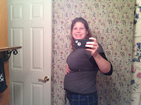 A smiling pregnant woman, roughly half-way through her pregnancy, taking a selfie and proudly holding her bump.  