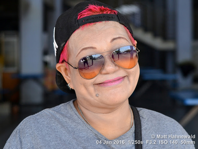 Close-up street portrait (outdoor headshot) of Cat, a remarkable Chinese-Malaysian lady from Kuah, wearing her signature baseball cup and distinctive make-up; Kuah, Langkawi, Malaysia. Matt Hahnewald Photography; Facing the World; closeup; street portrait; headshot; outdoor; Asia; Southeast Asia; Malaysia; Langkawi; Kuah; smiling; beautiful; Chinese Malaysian woman; red hair; cool; baseball cap; sunglasses; makeup