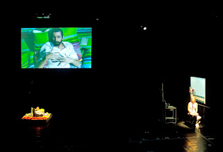 On the left of the stage, one actor is lying on a mattress, signing. A live cam projection shows his face and hands to the audience. On the right, an actress sits in a scenery representing a bathroom. She faces the audience and speaks into a microphone. The stage is dark except for spotlights on the actor and actress.