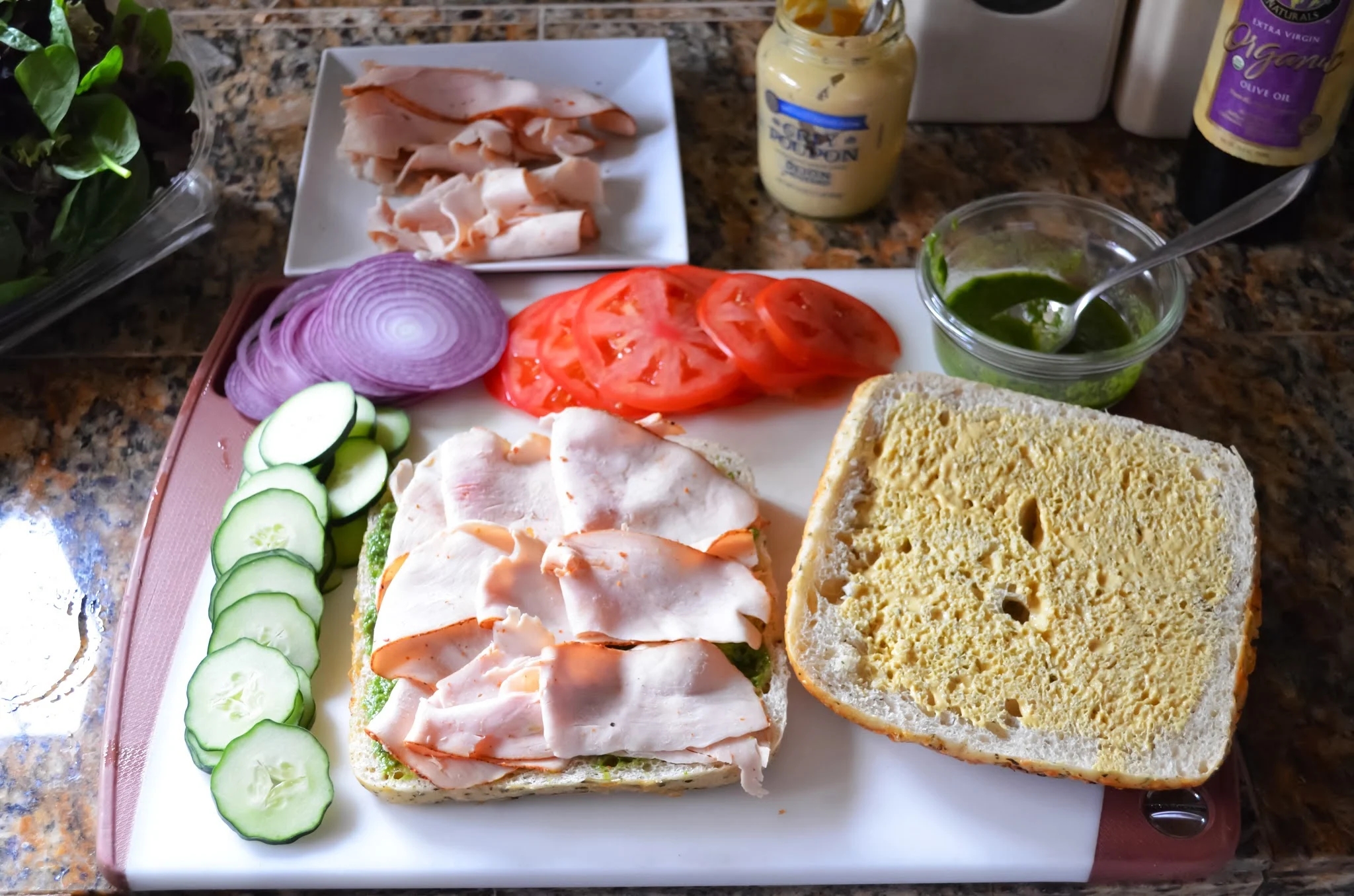 Turkey on top of pesto on focaccia bread with sliced cucumber, red onion and tomato on a white cutting board.