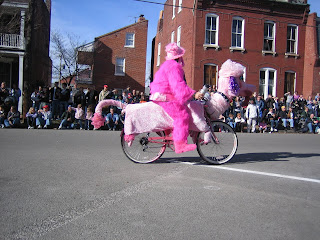 Woman in a pink furry costume riding a bicycle that looks like a pink dog.