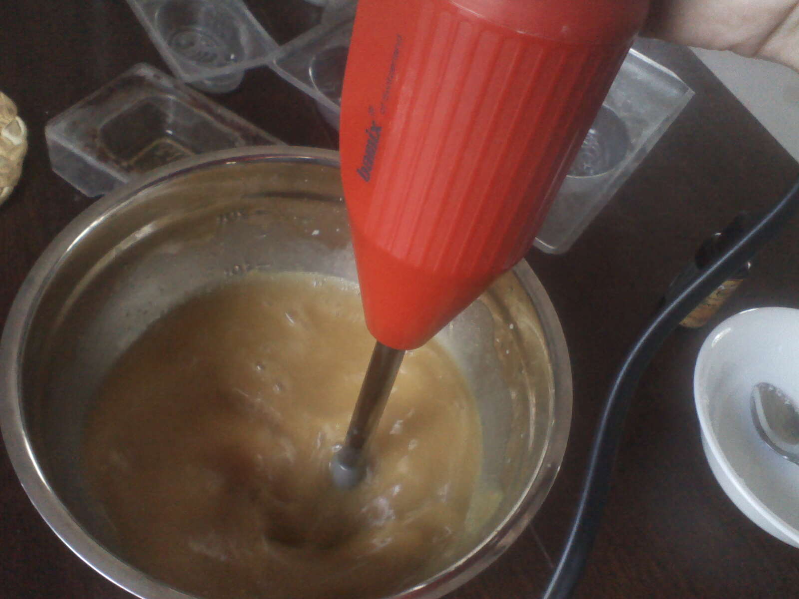 Image shows a large stock pot full of ingredients for homemade soap, with a stick blender immersed in the liquid, mixing it together.
