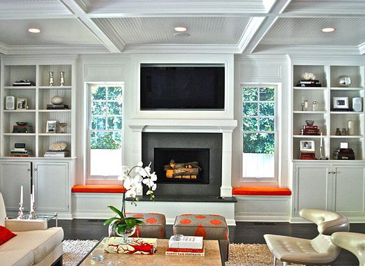 Den with coffered ceiling, built in bookshelves with windows seats with orange cushions, dark wood floor, a shag rug, two grey square stools with orange polka dots and two leather chairs with a white sofa