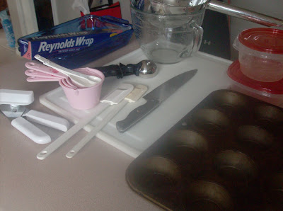 Measuring cups, knives, a cutting board, tin foil, plastic containers, and cupcake baking sheet, all ready to be used to prep for dinner.