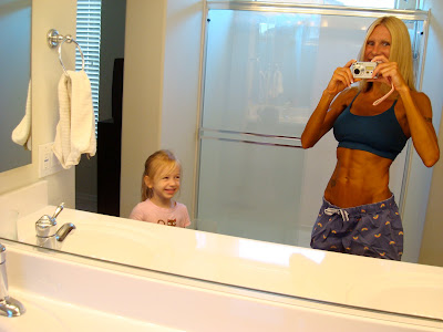 Woman in mirror showing off spray tan with young girl looking on