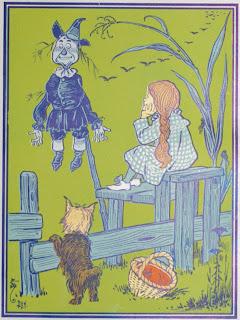 An illustration mostly in blue on a green backdrop, of a girl perched on a fence and looking at a scarecrow. A small dog also leans against the fence and there is a basket on the ground.