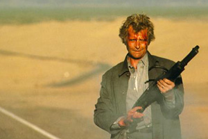 Um homem branco loiro (Rutger Hauer), com o rosto coberto de sangue, segura uma espingarda. Veste uma camisa cinza claro com uma jaqueta cinza por cima. Está parado em uma estrada rodeada com ambiente desértico.