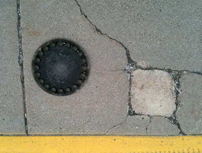 Round plastic hubcap near a square crack in a sidewalk, yellow curb at bottom