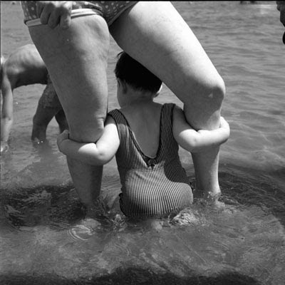 Child in bathing suit standing between adult woman's somewhat lumpy legs, grasping thighs