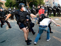 2 police officers in riot gear attack a single protester while a group of officers stand in the background also wearing riot gear