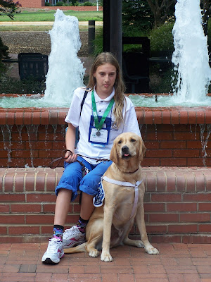 Photo of Brax & I. Brax is wearing a guide dog coat & in a sit-stay, I'm sitting down behind him