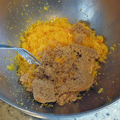 A close up photo of brown sugar being added to the orange zest and granulated sugar in a mixing bowl.