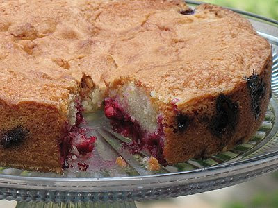 A close up photo of a cherry almond cake on a clear cake stand with slice missing .