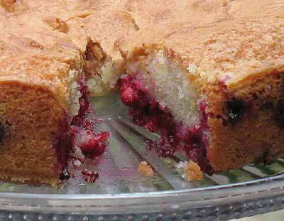 A close up photo of a cherry almond cake on a clear cake stand with slice missing .